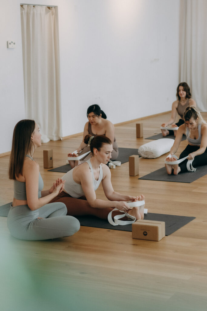 Yoga-Trainerin Mira,demonstriert eine sitzende Vorbeuge mit Gurt, während die Teilnehmerinnen ihr folgen.
