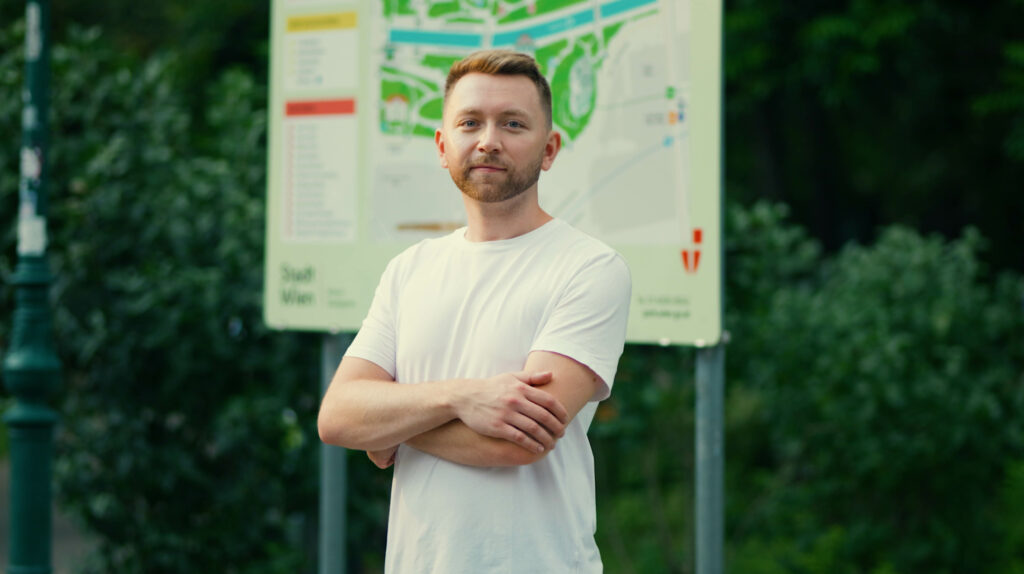 Ein Mann steht mit verschränkten Armen und selbstbewusstem Blick in die Kamera, im Hintergrund unscharf ein Parkplan.