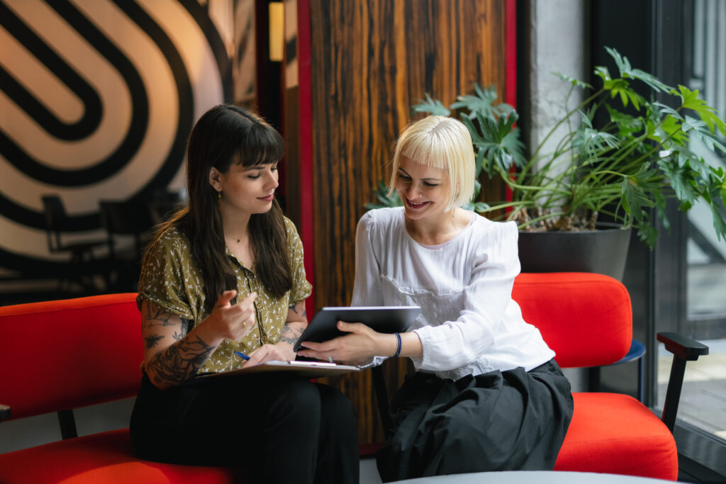 Zwei Frauen sitzen nebeneinander auf einem roten Sofa, eine mit Tablet, die andere mit Stift und Notizblock. Sie lächeln und sprechen miteinander, im Hintergrund Pflanzen und modernes Wanddesign.