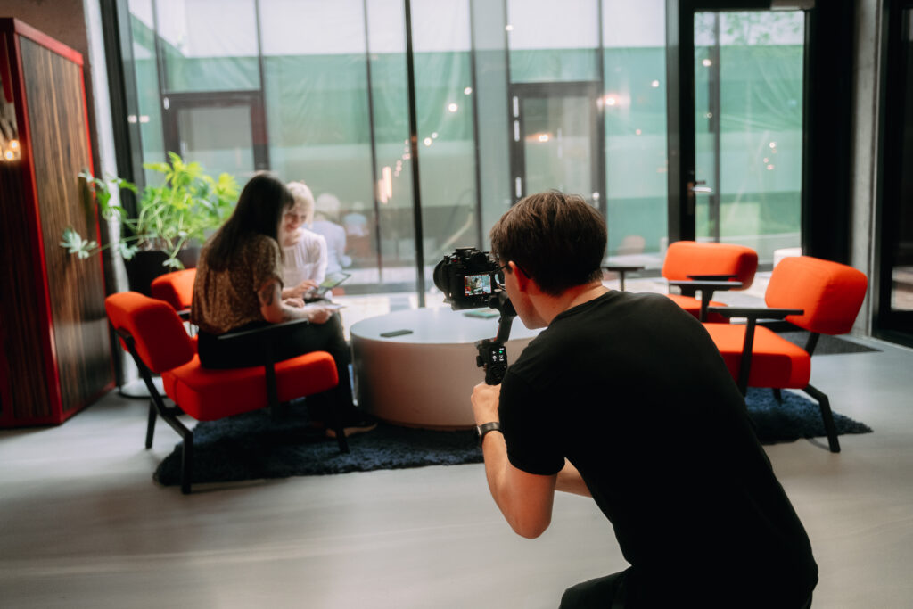 Kameramann Stephan kniet im Vordergrund mit einer Filmkamera auf einem Gimbal und filmt zwei Frauen, die sich an einem runden Tisch in roten Sesseln unterhalten. Im Hintergrund große Fensterfronten und moderne Büroeinrichtung. Behind The Scenes Einblicke von Pamina Films.